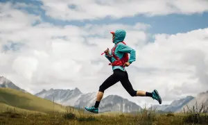 Kobieta w sportowym stroju i z plecakiem biegnie przez łąkę w górach.