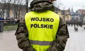 Żołnierz w kamizelce z napisem WOJSKO POLSKIE, w tle inni żołnierze i budynki. Skrót kapral widoczny na mundurze.