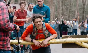 Zmęczona biegaczka opiera się o barierkę podczas Barkley Marathon. Tłum kibicuje, widząc jej determinację.