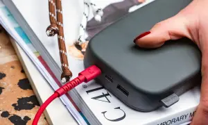 Dłoń z czerwonym lakierem na paznokciach dotyka szarego powerbanku. Czerwony kabel USB jest podłączony, co sugeruje, że trwa ładowanie.