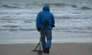 Mężczyzna w niebieskiej kurtce idzie po plaży z wykrywaczem metali i łopatą. Czy można chodzić z wykrywaczem po plaży? Tak, jeśli nie przeszkadza się innym.