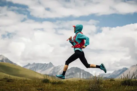 Kobieta w sportowym stroju i z plecakiem biegnie przez łąkę w górach.
