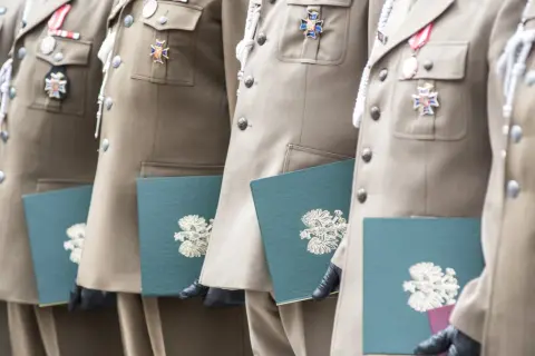 Żołnierze w mundurach z medalami odbierają dokumenty. Stare stopnie wojskowe zdobią ich piersi, a w rękach trzymają zielone teczki z godłem.