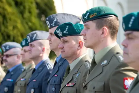 Żołnierze w różnych rodzajach mundurów wojskowych, w tym w zielonych beretach i szarych furażerkach, prezentują swoje odznaczenia i stopnie wojskowe.