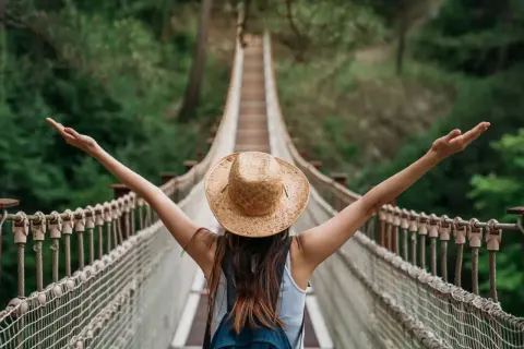 Kobieta w kapeluszu na moście wiszącym, z ramionami rozłożonymi w geście wolności. Przestrzega zasad "leave no trace", ciesząc się naturą.