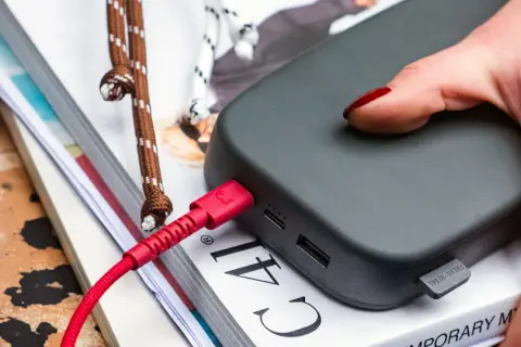 Dłoń z czerwonym lakierem na paznokciach dotyka szarego powerbanku. Czerwony kabel USB jest podłączony, co sugeruje, że trwa ładowanie.