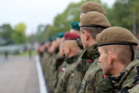 Żołnierze w kamuflażu i beretach stoją w szeregu. To kategoria d wojsko.