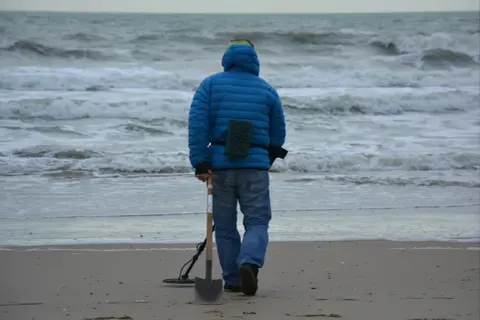 Mężczyzna w niebieskiej kurtce idzie po plaży z wykrywaczem metali i łopatą. Czy można chodzić z wykrywaczem po plaży? Tak, jeśli nie przeszkadza się innym.