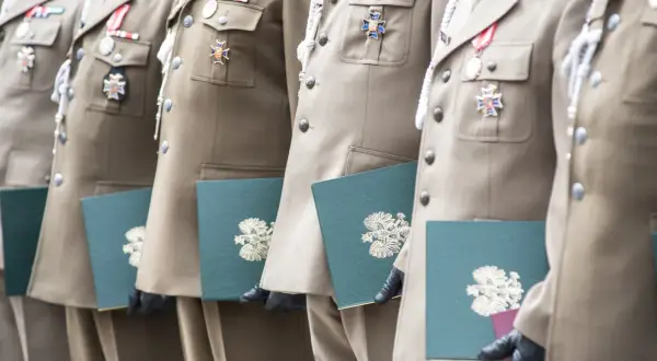 Żołnierze w mundurach z medalami odbierają dokumenty. Stare stopnie wojskowe zdobią ich piersi, a w rękach trzymają zielone teczki z godłem.