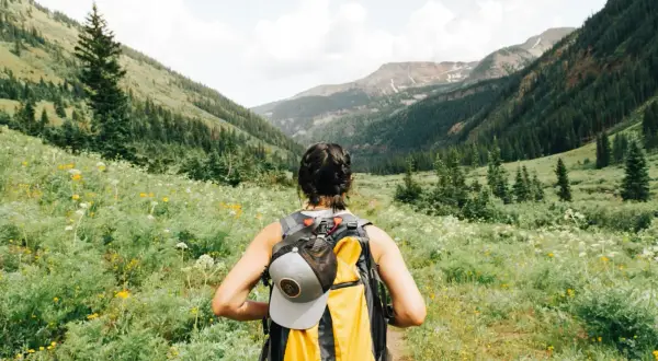 Kobieta z plecakiem wyrusza na trekking przez kwiecistą dolinę, podziwiając górskie krajobrazy.