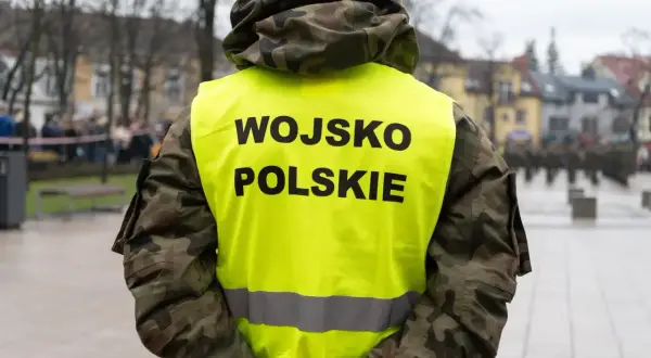 Żołnierz w kamizelce z napisem WOJSKO POLSKIE, w tle inni żołnierze i budynki. Skrót kapral widoczny na mundurze.