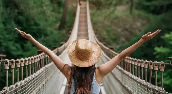 Kobieta w kapeluszu na moście wiszącym, z ramionami rozłożonymi w geście wolności. Przestrzega zasad "leave no trace", ciesząc się naturą.