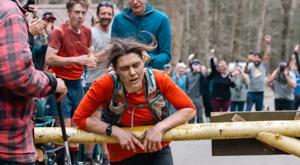 Zmęczona biegaczka opiera się o barierkę podczas Barkley Marathon. Tłum kibicuje, widząc jej determinację.