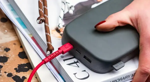 Dłoń z czerwonym lakierem na paznokciach dotyka szarego powerbanku. Czerwony kabel USB jest podłączony, co sugeruje, że trwa ładowanie.