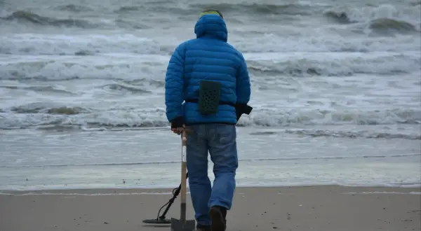Mężczyzna w niebieskiej kurtce idzie po plaży z wykrywaczem metali i łopatą. Czy można chodzić z wykrywaczem po plaży? Tak, jeśli nie przeszkadza się innym.
