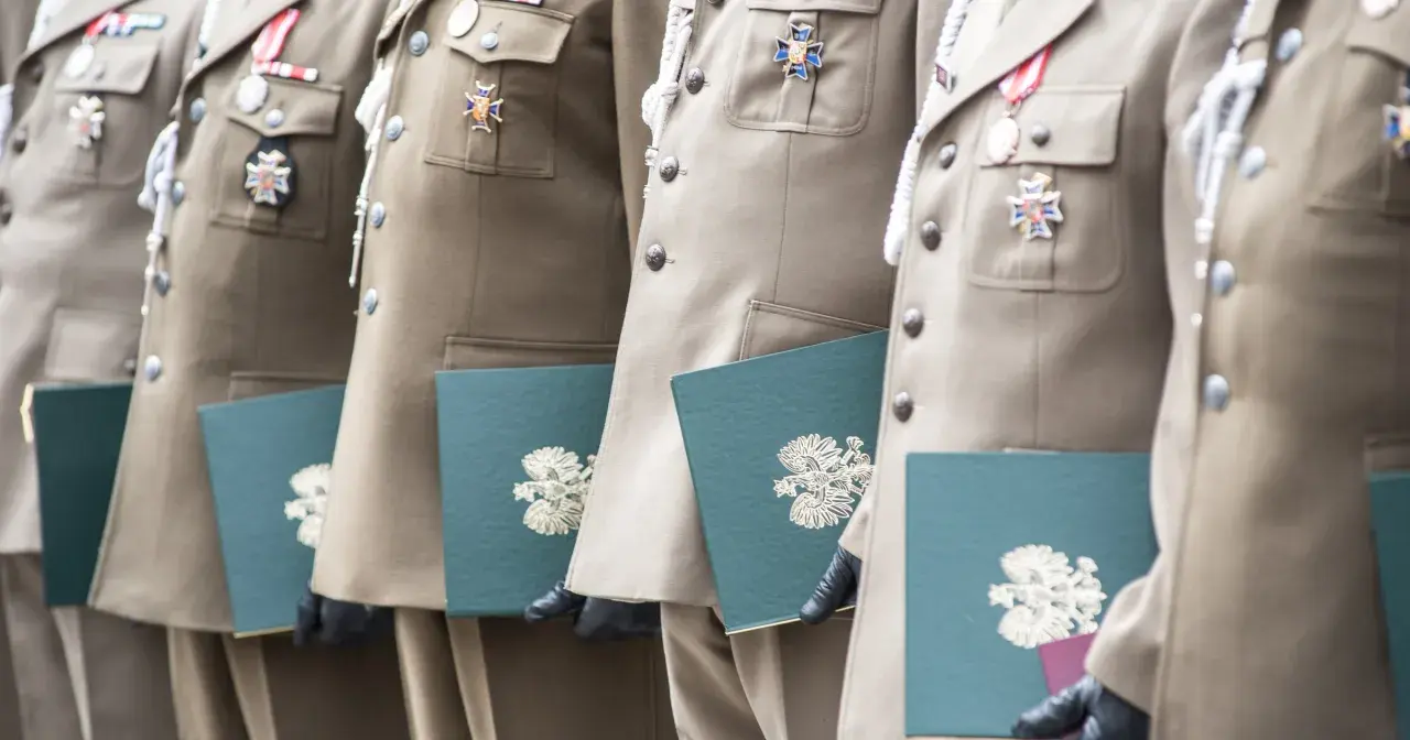 Żołnierze w mundurach z medalami odbierają dokumenty. Stare stopnie wojskowe zdobią ich piersi, a w rękach trzymają zielone teczki z godłem.