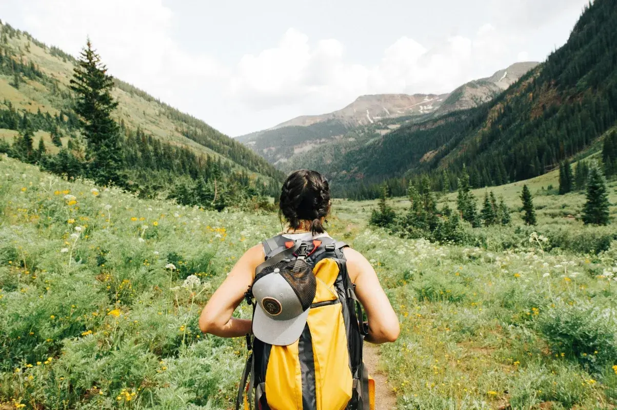 Kobieta z plecakiem wyrusza na trekking przez kwiecistą dolinę, podziwiając górskie krajobrazy.