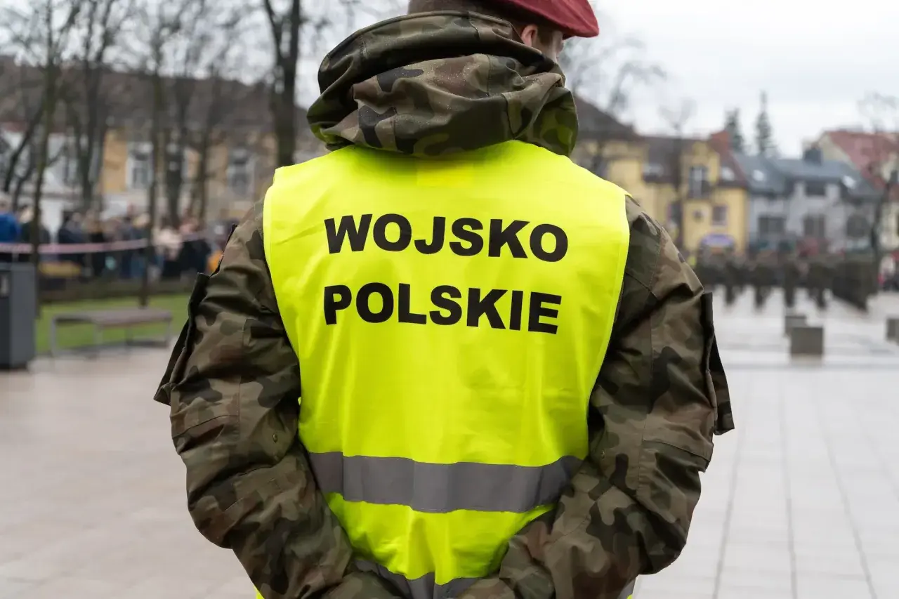 Żołnierz w kamizelce z napisem WOJSKO POLSKIE, w tle inni żołnierze i budynki. Skrót kapral widoczny na mundurze.