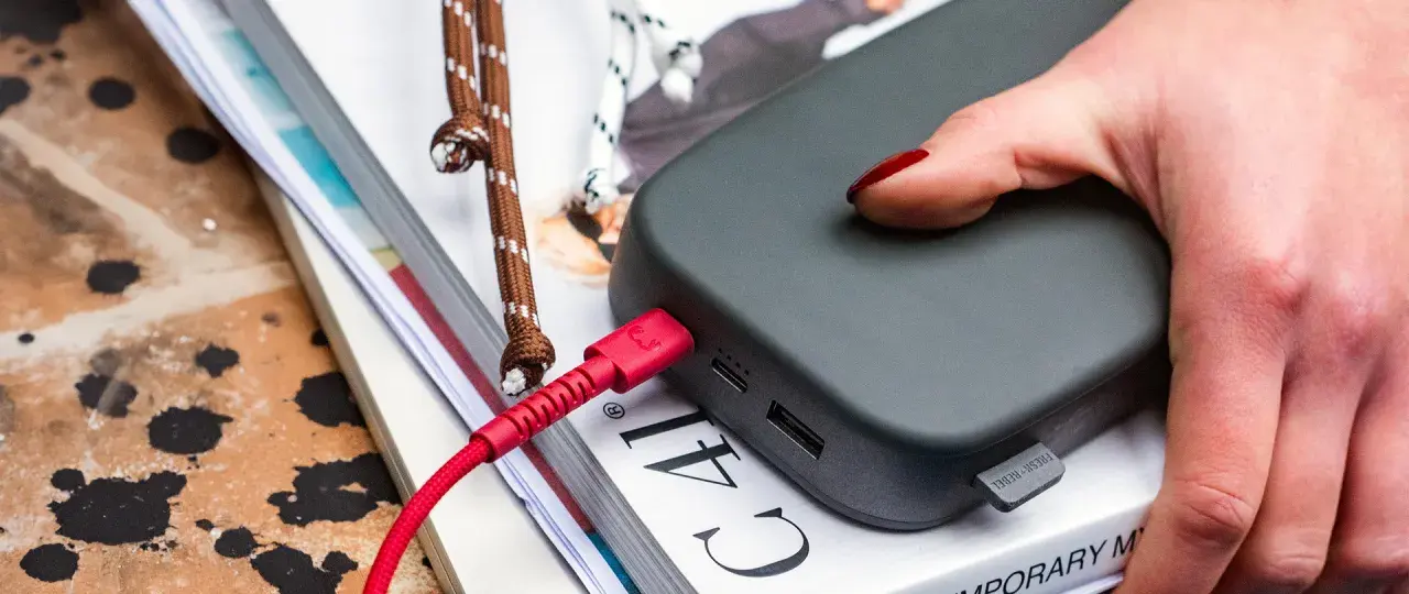 Dłoń z czerwonym lakierem na paznokciach dotyka szarego powerbanku. Czerwony kabel USB jest podłączony, co sugeruje, że trwa ładowanie.
