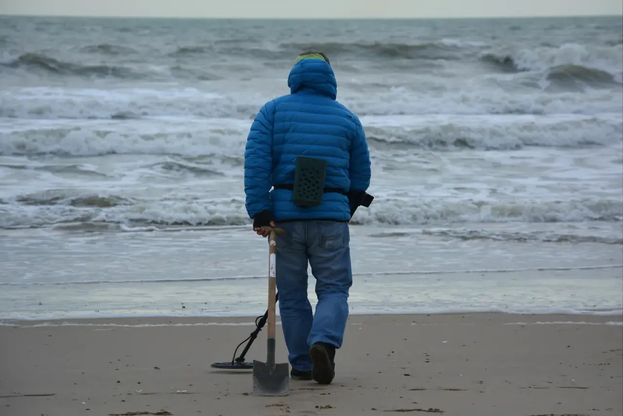 Mężczyzna w niebieskiej kurtce idzie po plaży z wykrywaczem metali i łopatą. Czy można chodzić z wykrywaczem po plaży? Tak, jeśli nie przeszkadza się innym.