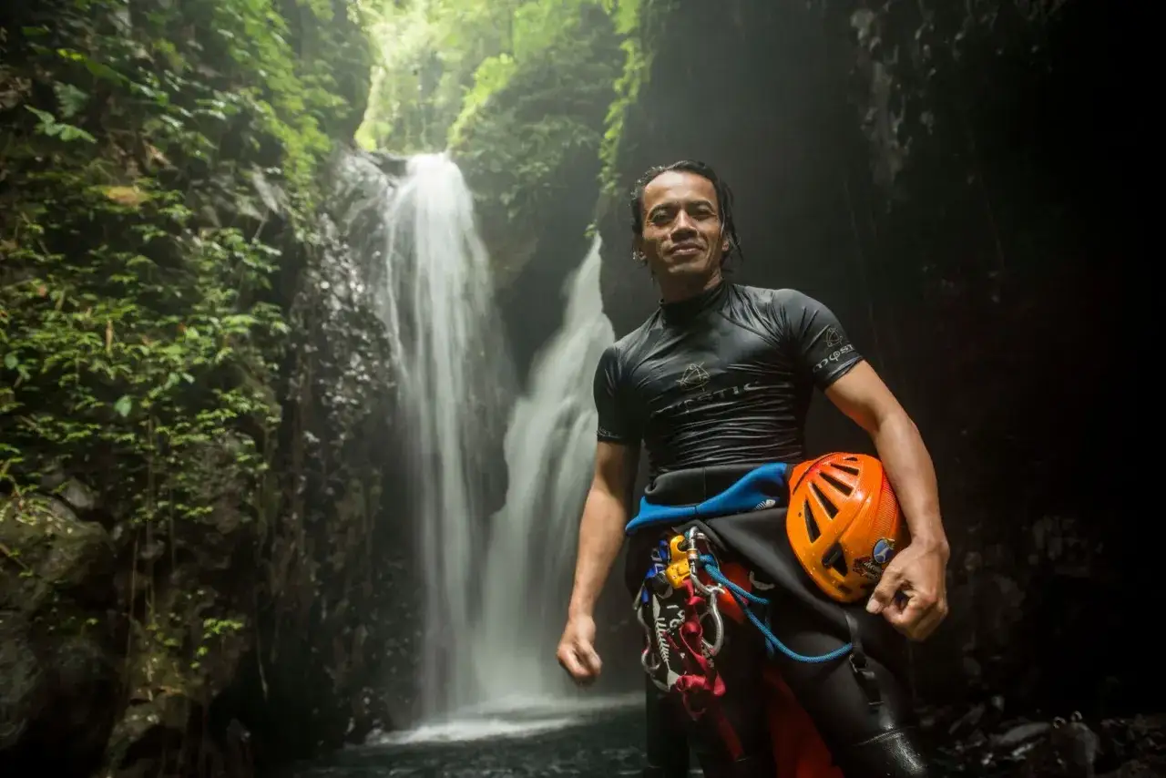 Mężczyzna w stroju do canyoningu z kaskiem w ręku, obok wodospadu. To właśnie jest canyoning, co to za przygoda!