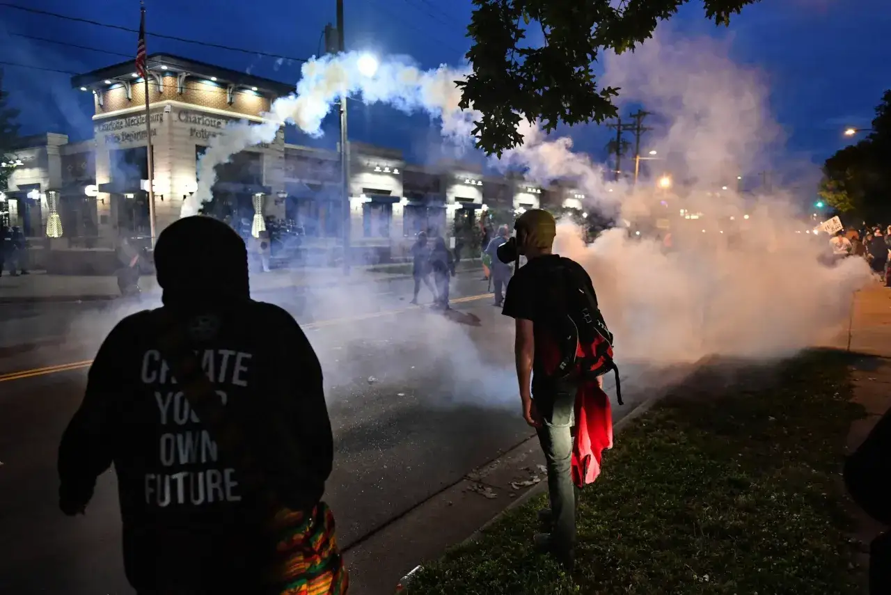 Dym z gazu pieprzowego unosi się przed budynkiem policji. Ludzie w maskach i z transparentami stoją w kłębach dymu, doświadczając jego skutków.