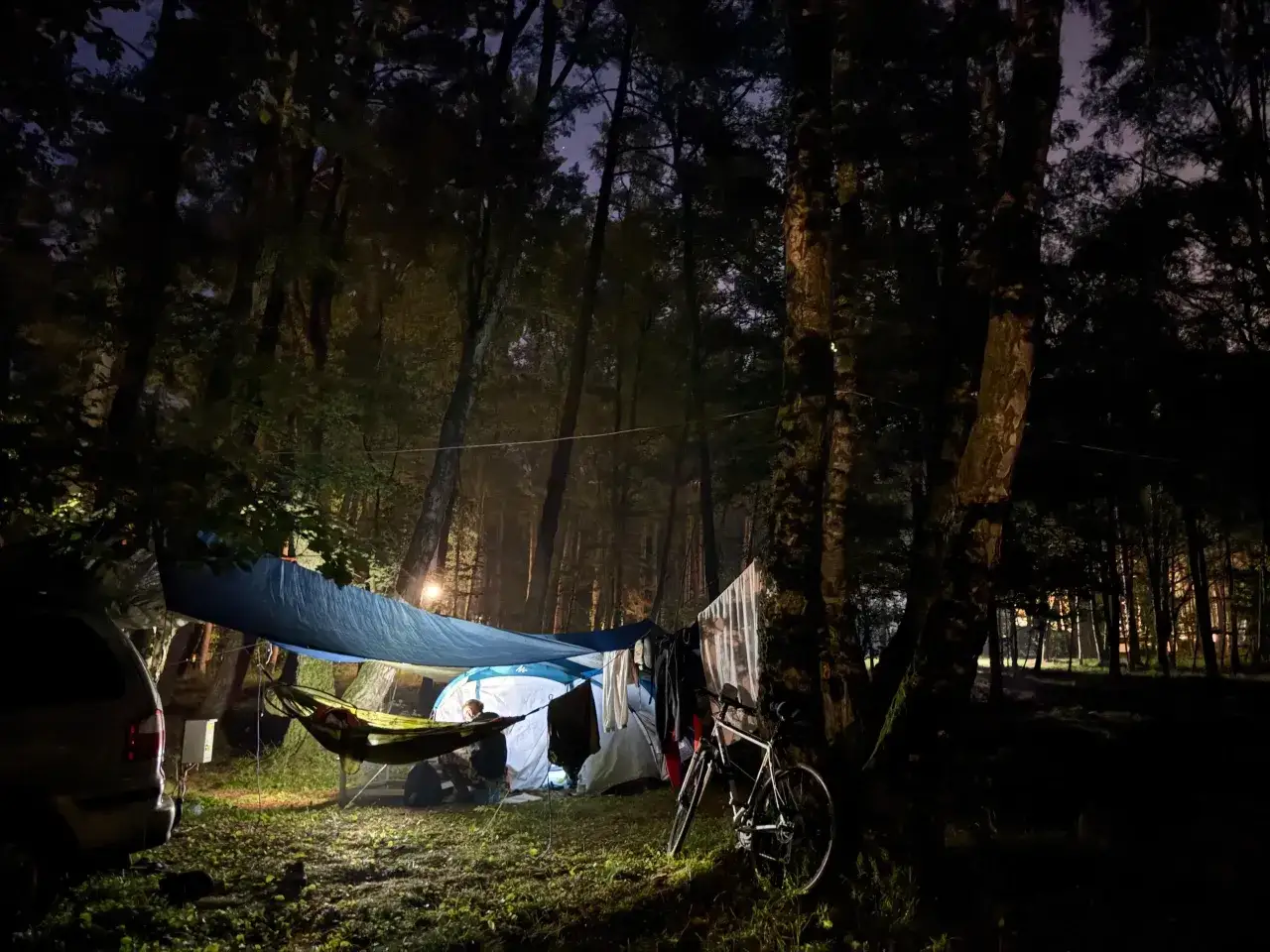 Nocne spanie pod namiotem w lesie. Rozstawiony namiot, hamak, rower i samoch&oacute;d tworzą obozowisko.