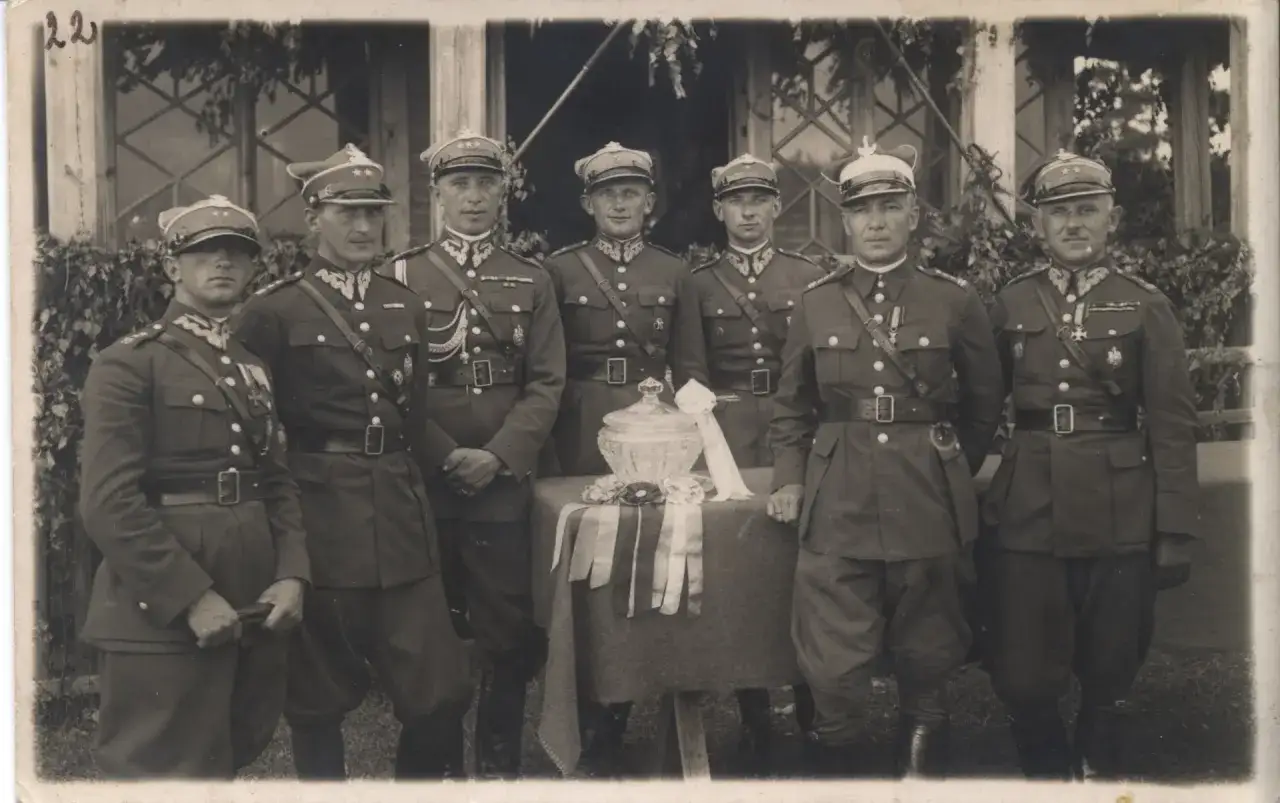 Siedmiu mężczyzn w mundurach wojskowych, wśr&oacute;d nich rotmistrz odpowiednik stopnia, pozuje do zdjęcia przed budynkiem.