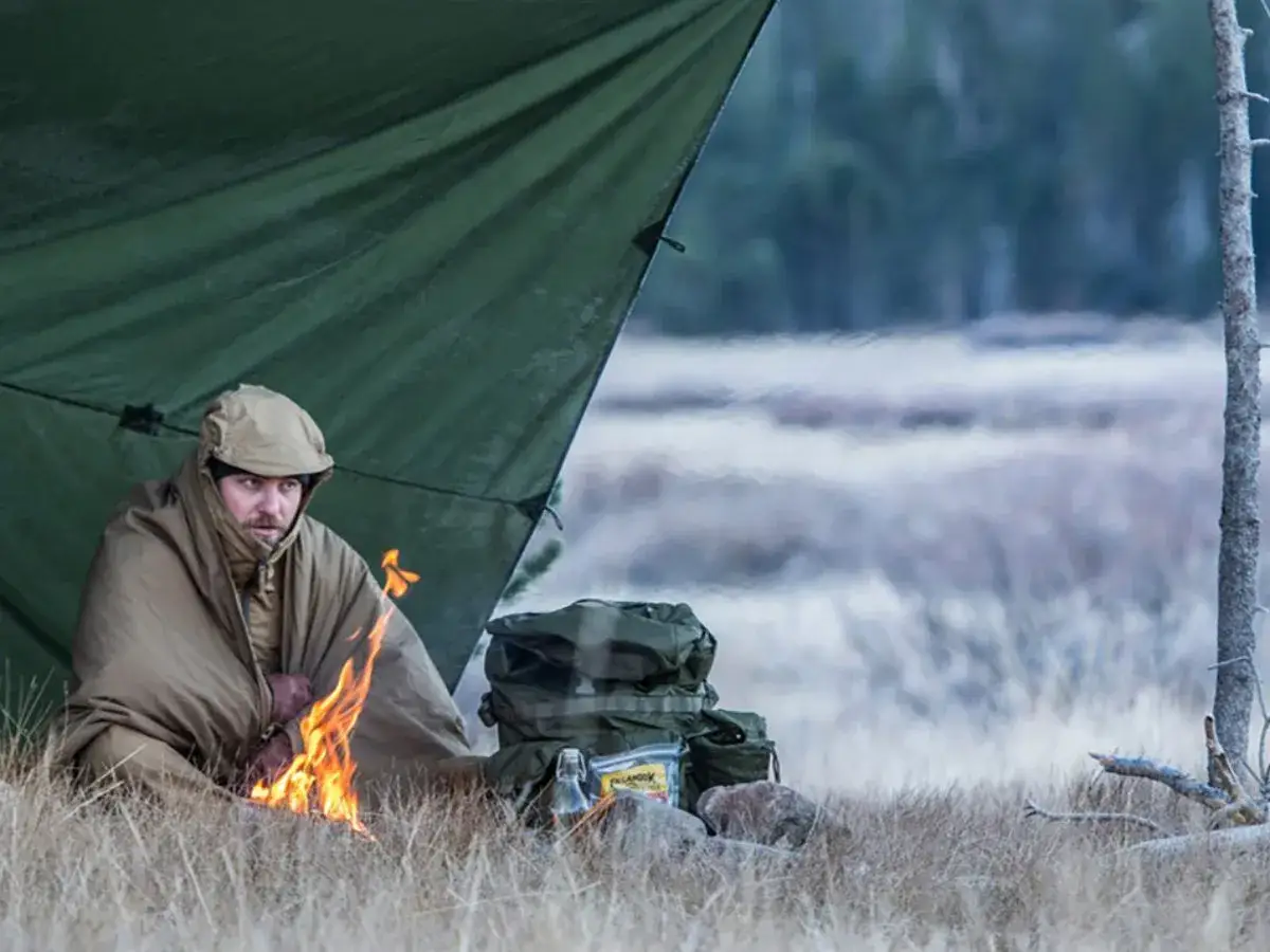 Mężczyzna w śpiworze przy ognisku pod tarpem, obok plecak i butelka. Podstawowe zasady survivalu w dziczy.