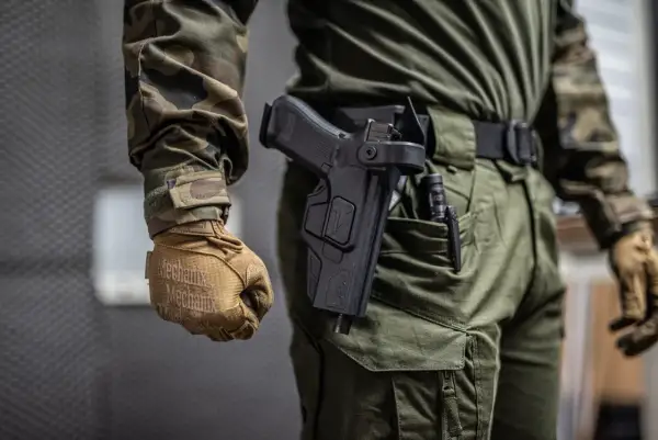 Osoba w mundurze taktycznym, z rękawiczkami Mechanix, prezentuje noszenie broni w kaburze na pasie.