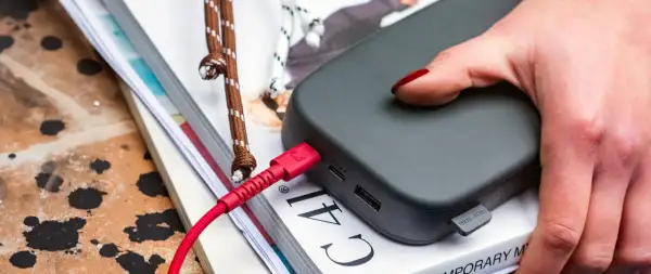 Dłoń z czerwonym lakierem na paznokciach dotyka szarego powerbanku. Czerwony kabel USB jest podłączony, co sugeruje, że trwa ładowanie.
