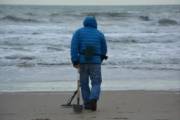 Mężczyzna w niebieskiej kurtce idzie po plaży z wykrywaczem metali i łopatą. Czy można chodzić z wykrywaczem po plaży? Tak, jeśli nie przeszkadza się innym.